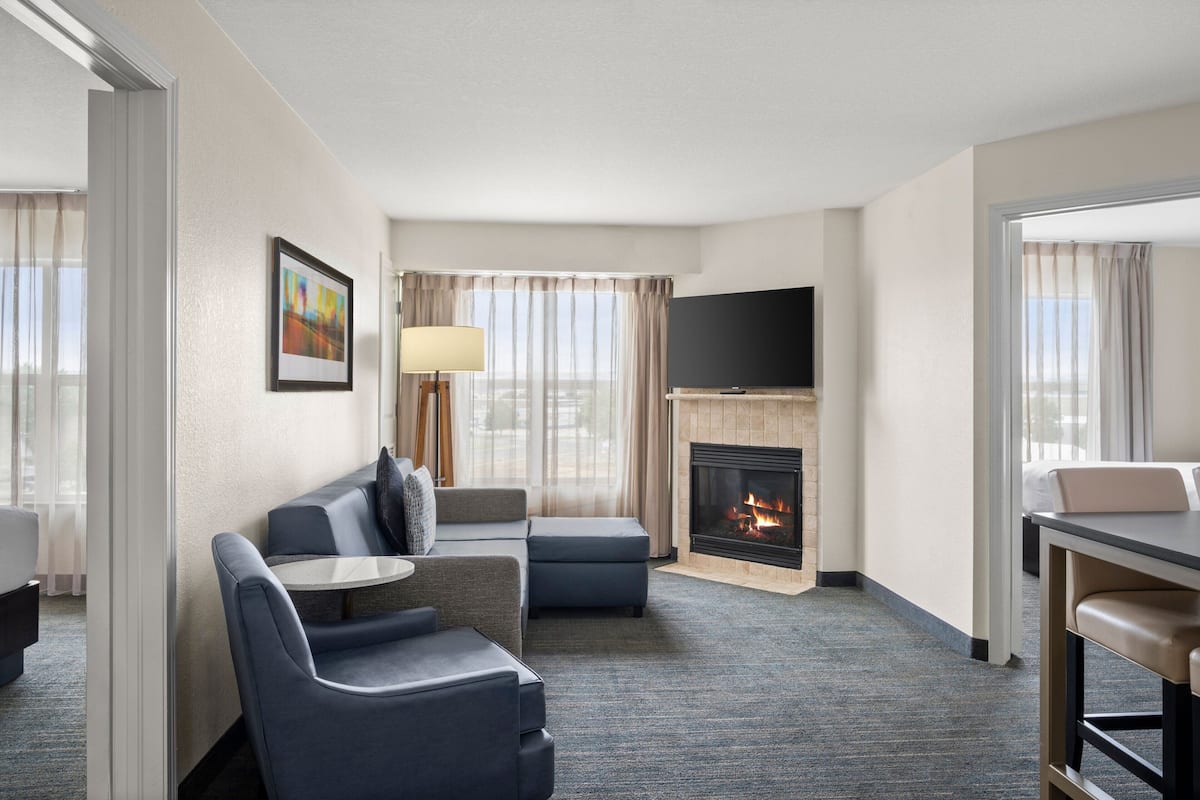 Habitación de hotel con sofá, sillón, reposapiés, televisor de pantalla plana, chimenea y mesa de comedor.
