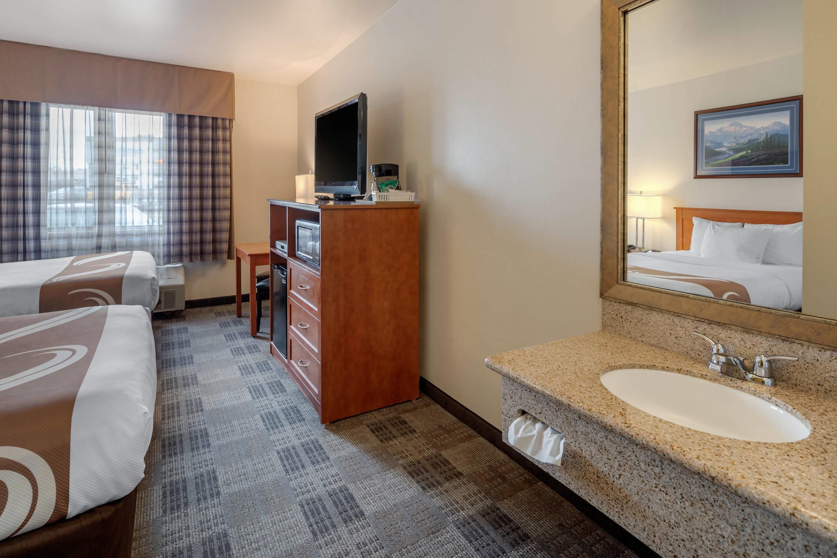 A hotel room with two beds, a television, a desk, a mirror, and a bathroom with a sink and mirror.