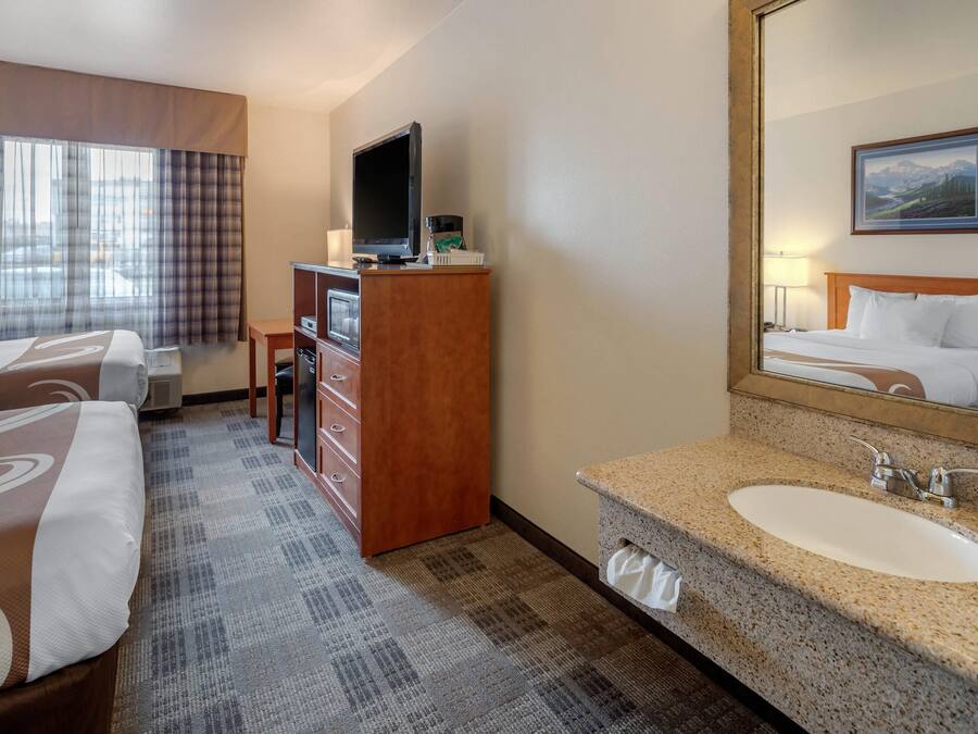 A hotel room with two beds, a television, a desk, a mirror, and a bathroom with a sink and mirror.