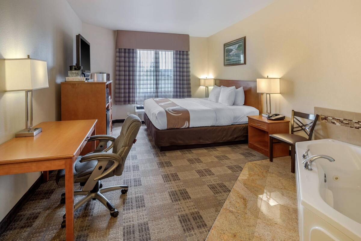 Habitación de hotel con cama, escritorio, silla, lámpara de escritorio, mesita de noche, lámpara y bañera.