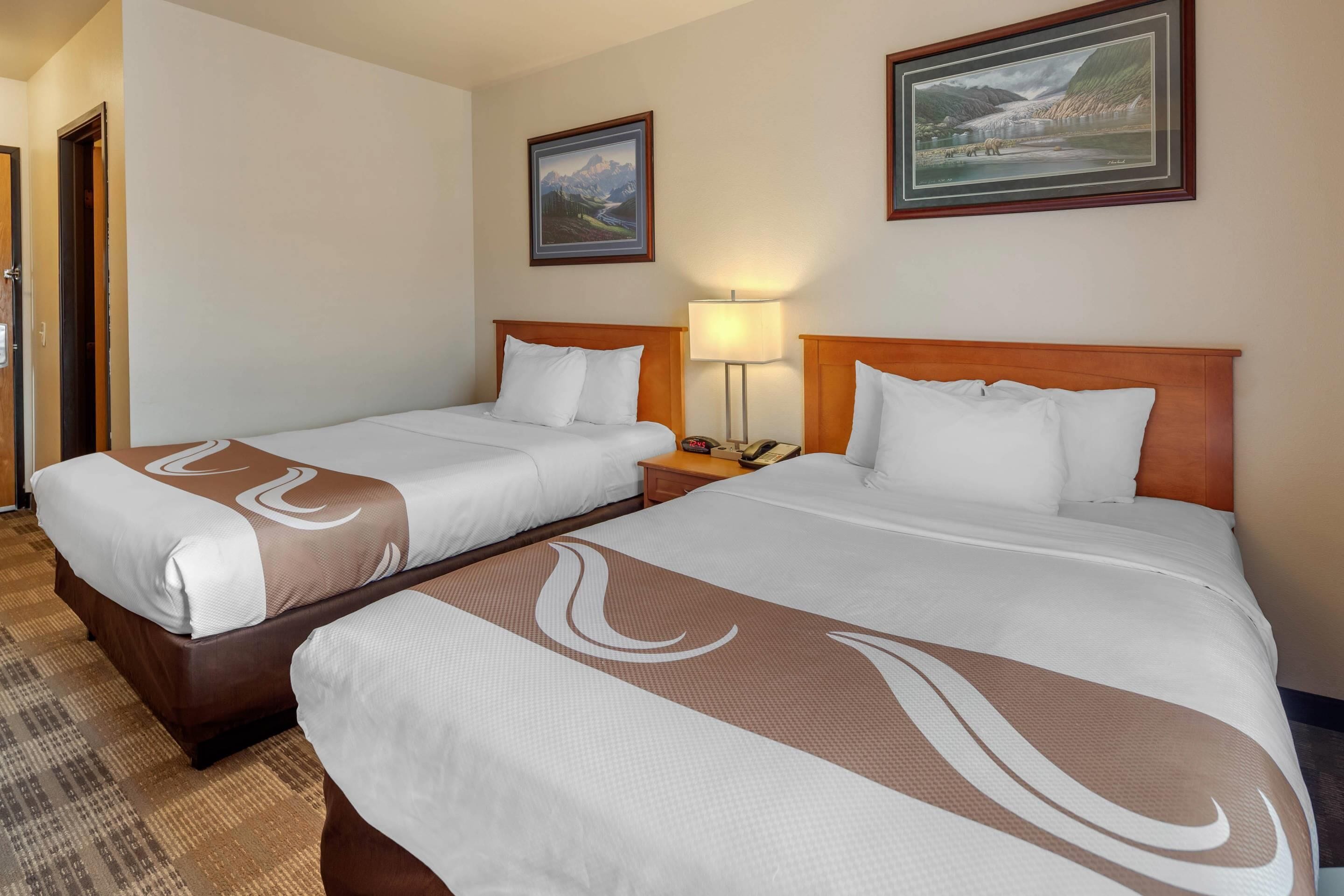 Una camera d'albergo con due letti, un comodino con una lampada e due quadri appesi al muro.