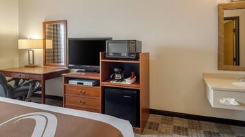 Habitación de hotel con cama, escritorio con lámpara, televisión, microondas, cafetera, refrigerador, espejo y baño con lavamanos y dispensador de toallas.