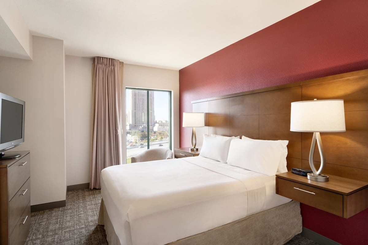 A hotel room with a large bed, two bedside tables with lamps, a television, a window with curtains, and a city view.
