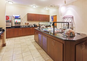 Un área de desayuno de hotel con un mostrador de autoservicio, un refrigerador de Coca-Cola y una máquina de café.