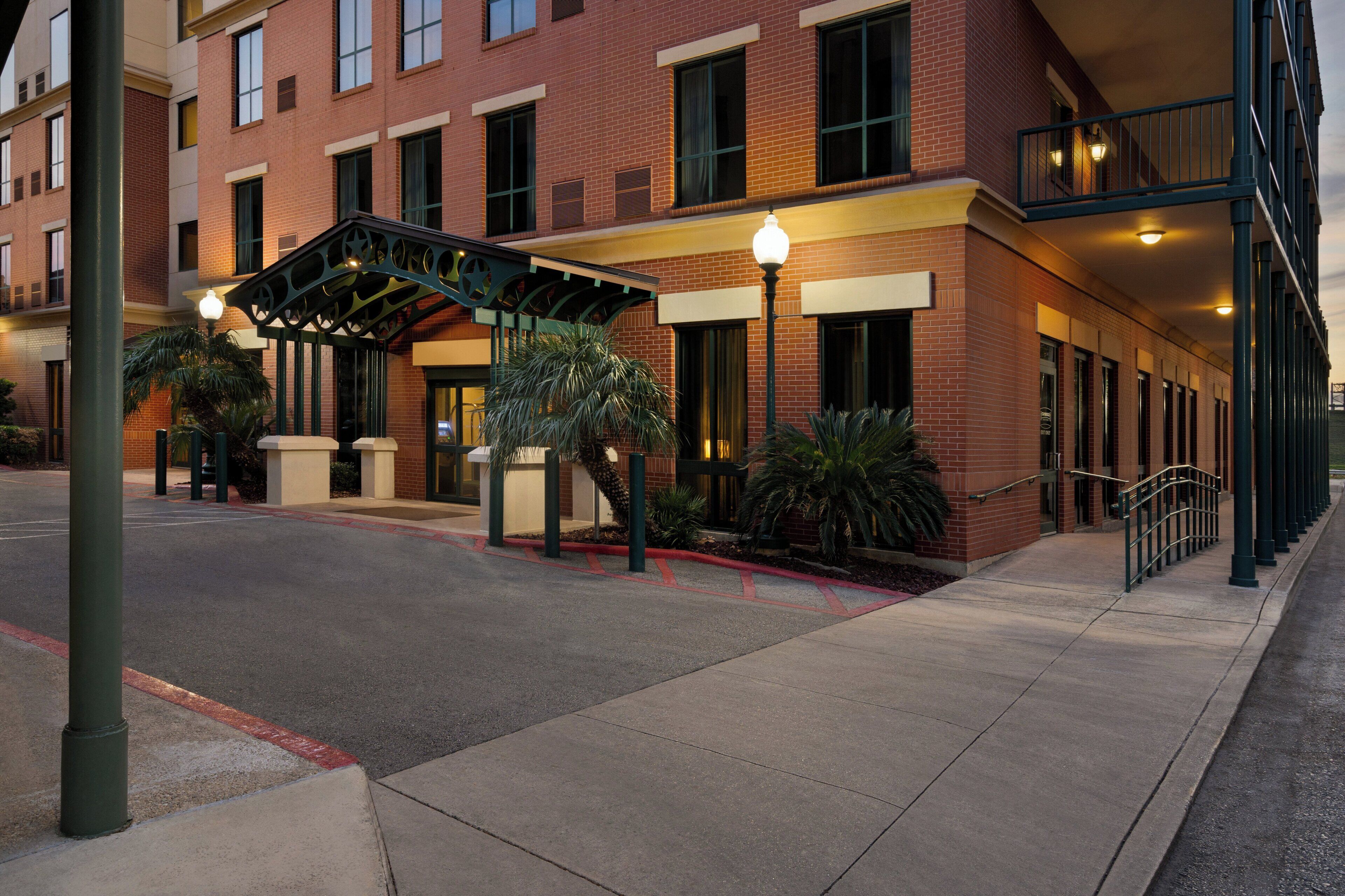 A hotel entrance with a covered walkway, a parking lot, and a building with multiple windows and a brick facade.