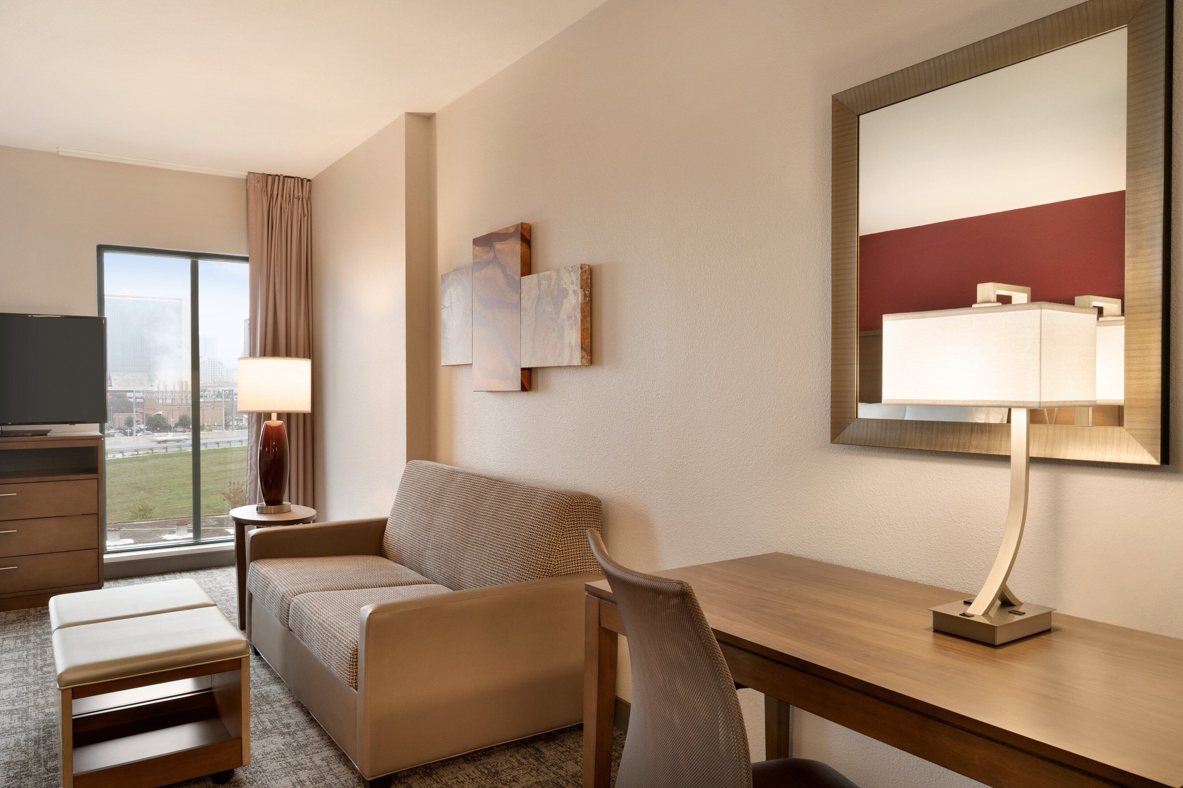Et hotelværelse med sofa, skrivebord, lampe, fjernsyn og et stort vindue med byudsigt.
