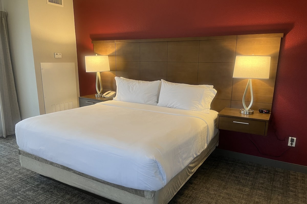A hotel room with a large bed, two bedside tables with lamps, a nightstand, a wardrobe, and a thermostat on the wall.