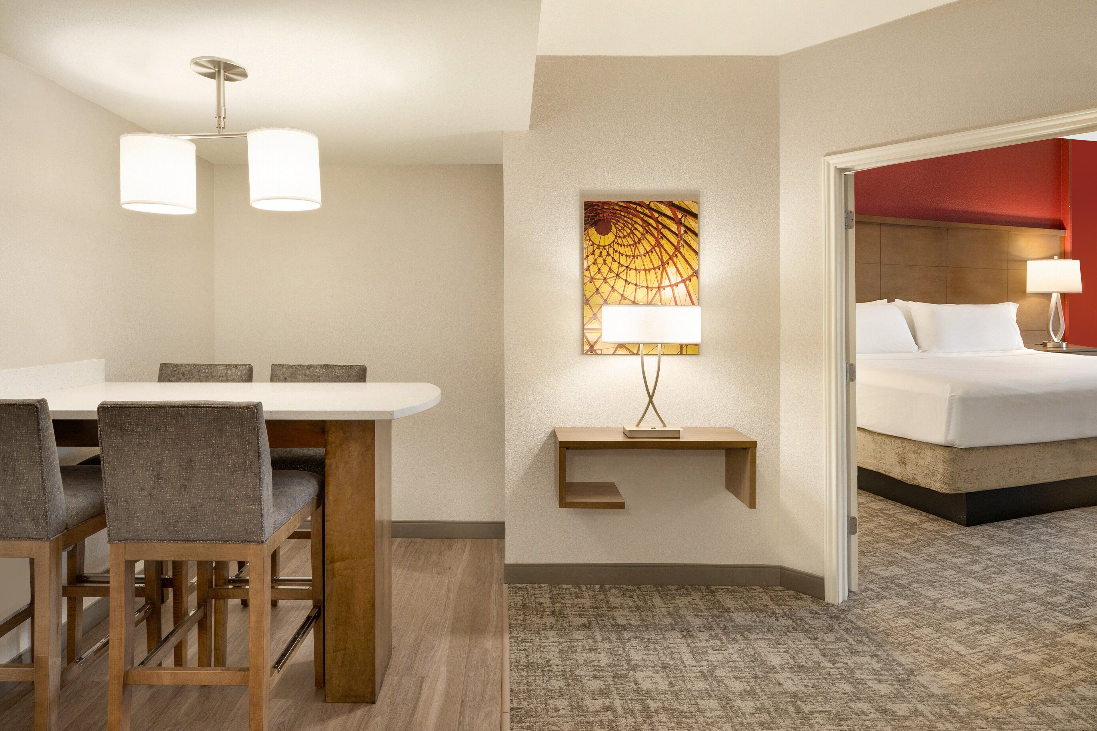 A hotel room with a bed, a desk with chairs, a lamp, and a painting on the wall.