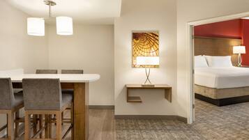 A hotel room with a bed, a desk with chairs, a lamp, and a painting on the wall.