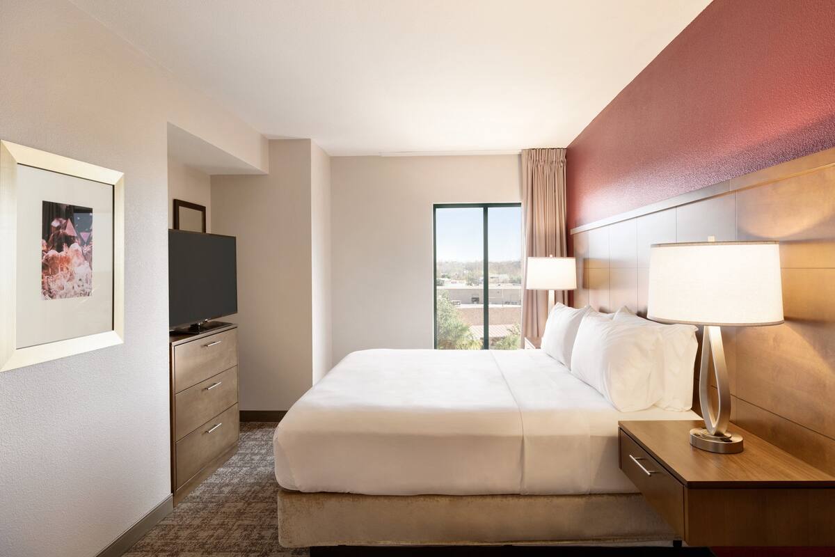 A hotel room with a large bed, a nightstand with a lamp, a TV on a stand, a dresser, and a window with a view of the city.