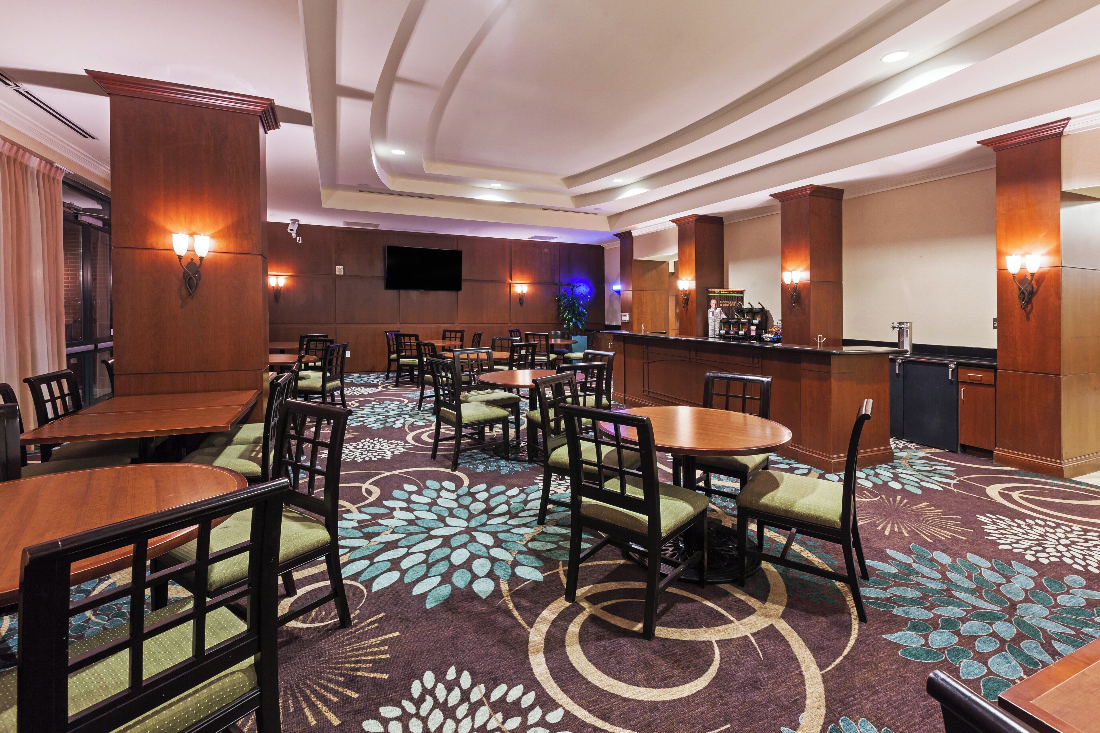 A hotel dining area with multiple tables and chairs, a bar, and a flat-screen TV mounted on the wall.