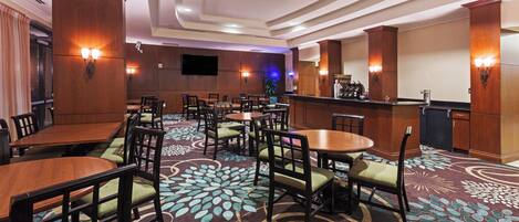 A hotel dining area with multiple tables and chairs, a bar, and a flat-screen TV mounted on the wall.