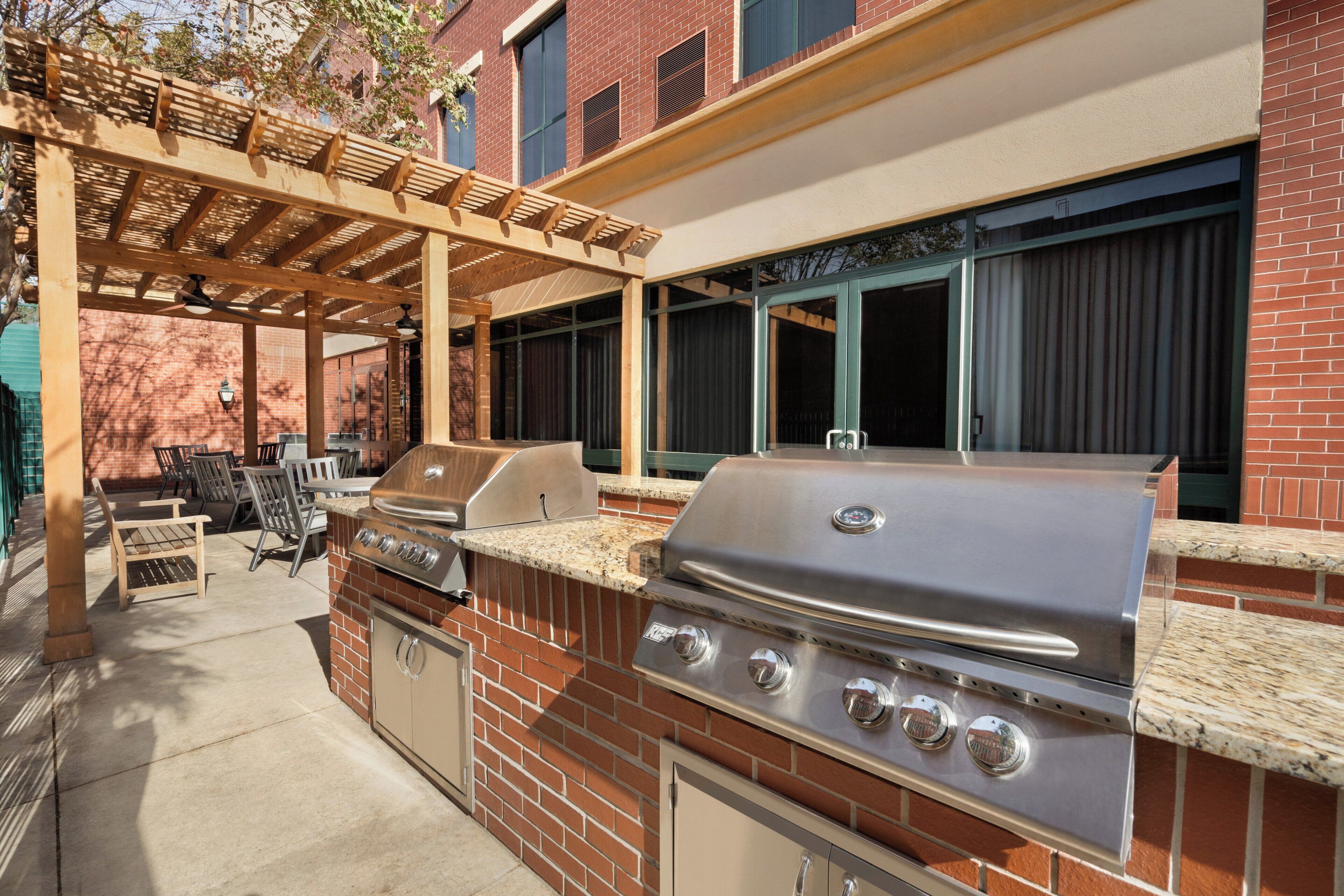 An outdoor patio with a brick barbecue grill, wooden chairs, and a pergola.
