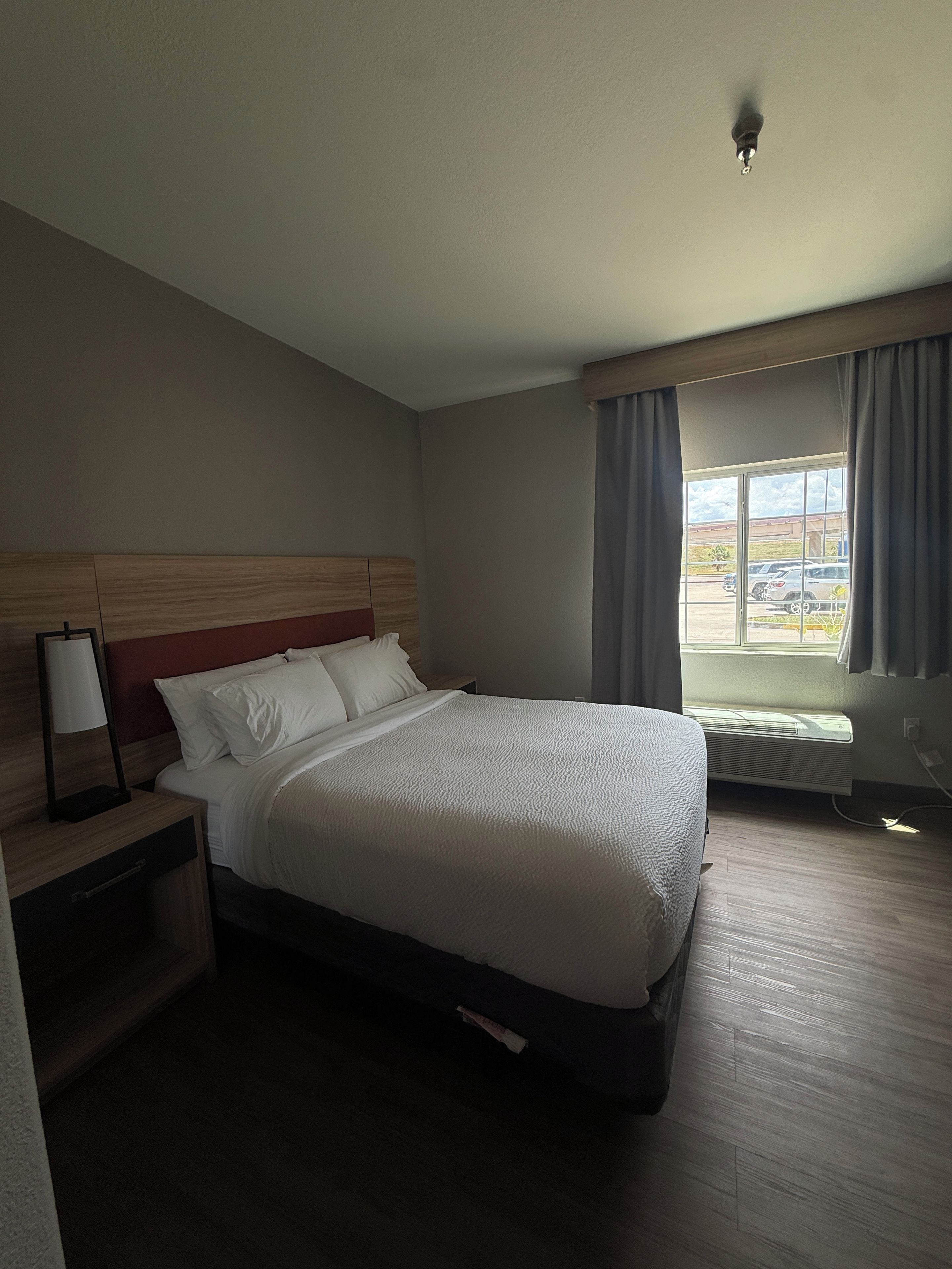 Ein Hotelzimmer mit Bett, Nachttisch, Lampe, einem Fenster mit Vorhängen und einer Klimaanlage.