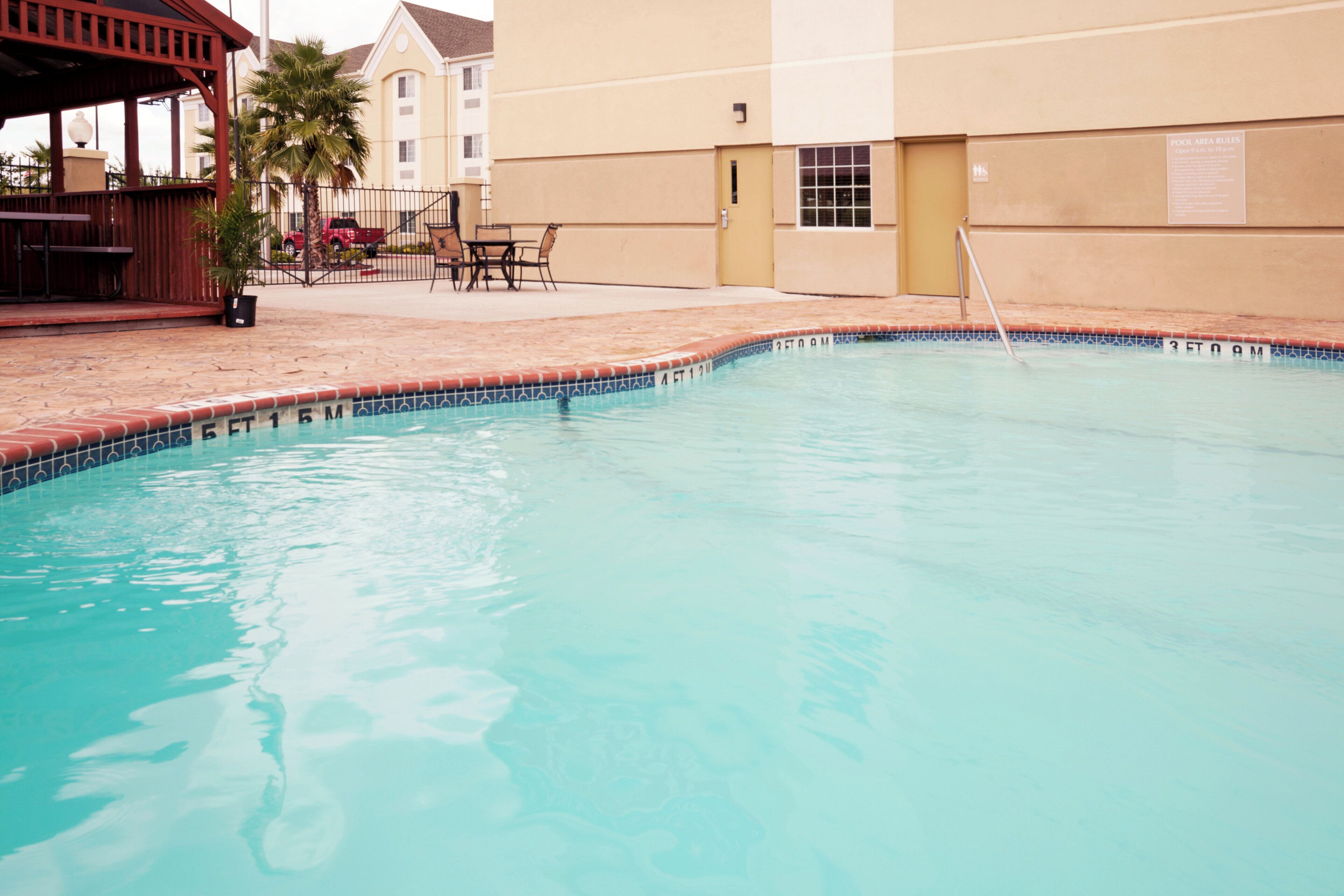 A swimming pool with a tiled edge, a lounge area with outdoor seating, and a building in the background.