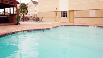 A swimming pool with a tiled edge, a lounge area with outdoor seating, and a building in the background.