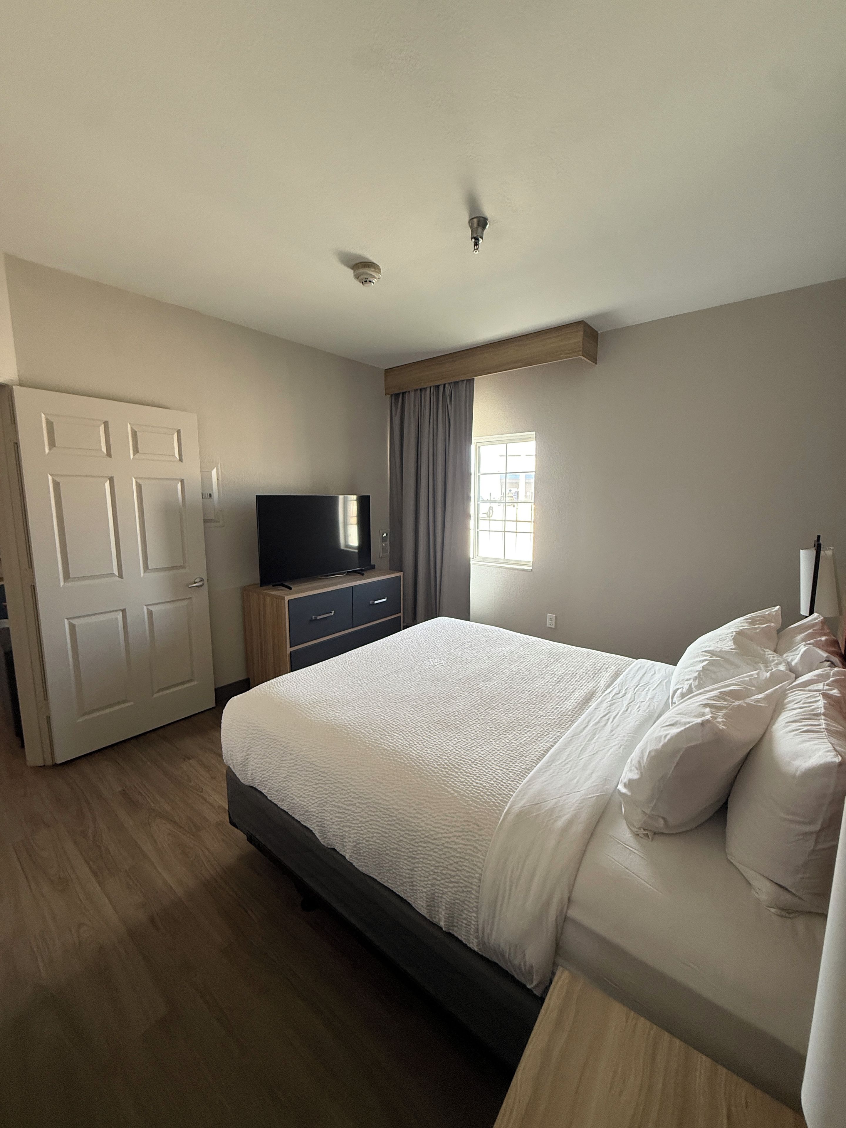 Une chambre à coucher avec un lit, un téléviseur posé sur une commode en bois, une fenêtre avec des rideaux et une porte.