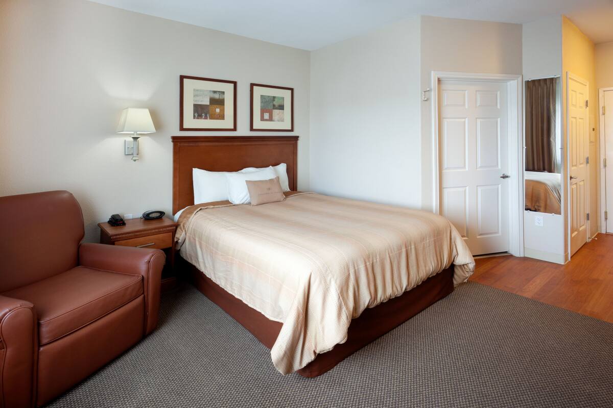 A hotel room with a bed, a brown leather chair, a nightstand, a lamp, and two framed pictures on the wall.