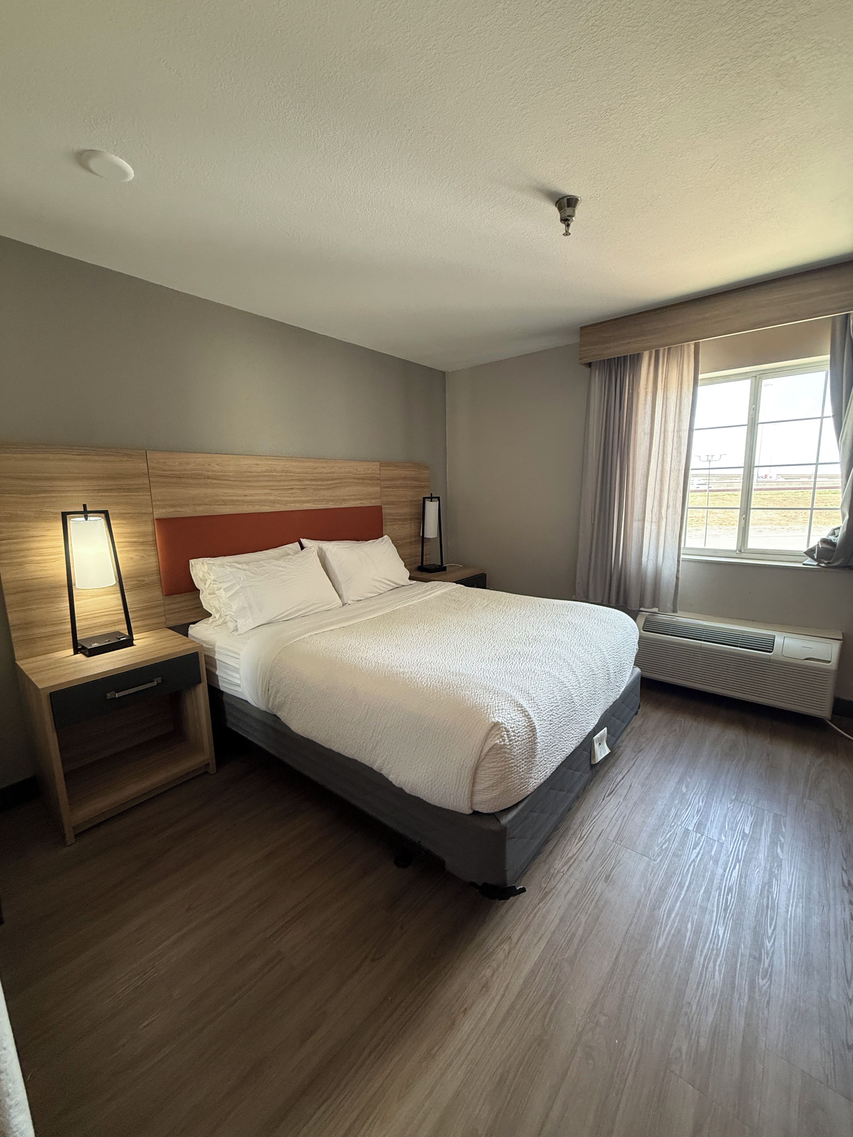 Una habitación de hotel con cama, mesita de noche, lámpara, ventana con cortinas y radiador.