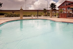 Una piscina con borde de azulejos, rodeada por una cerca y una zona cubierta a la derecha.
