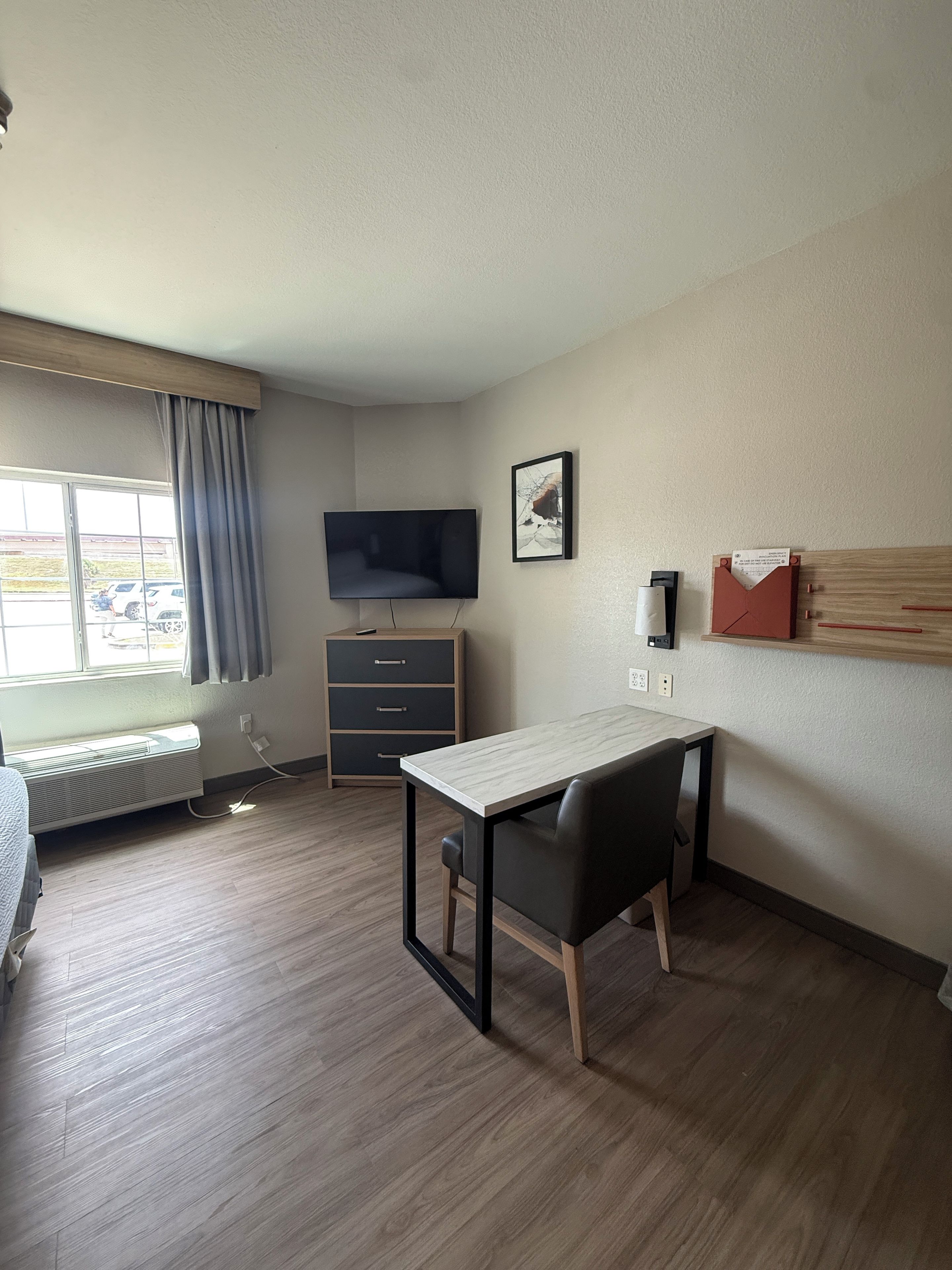 A hotel room with a bed, a desk with a chair, a television, a window with curtains, a heater, and a wall-mounted shelf.