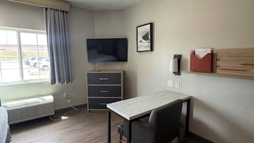 A hotel room with a bed, a desk with a chair, a television, a window with curtains, a heater, and a wall-mounted shelf.