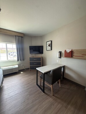 A hotel room with a bed, a desk with a chair, a television, a window with curtains, a heater, and a wall-mounted shelf. - Candlewood Suites Corpus Christi-Spid by IHG (Corpus Christi)