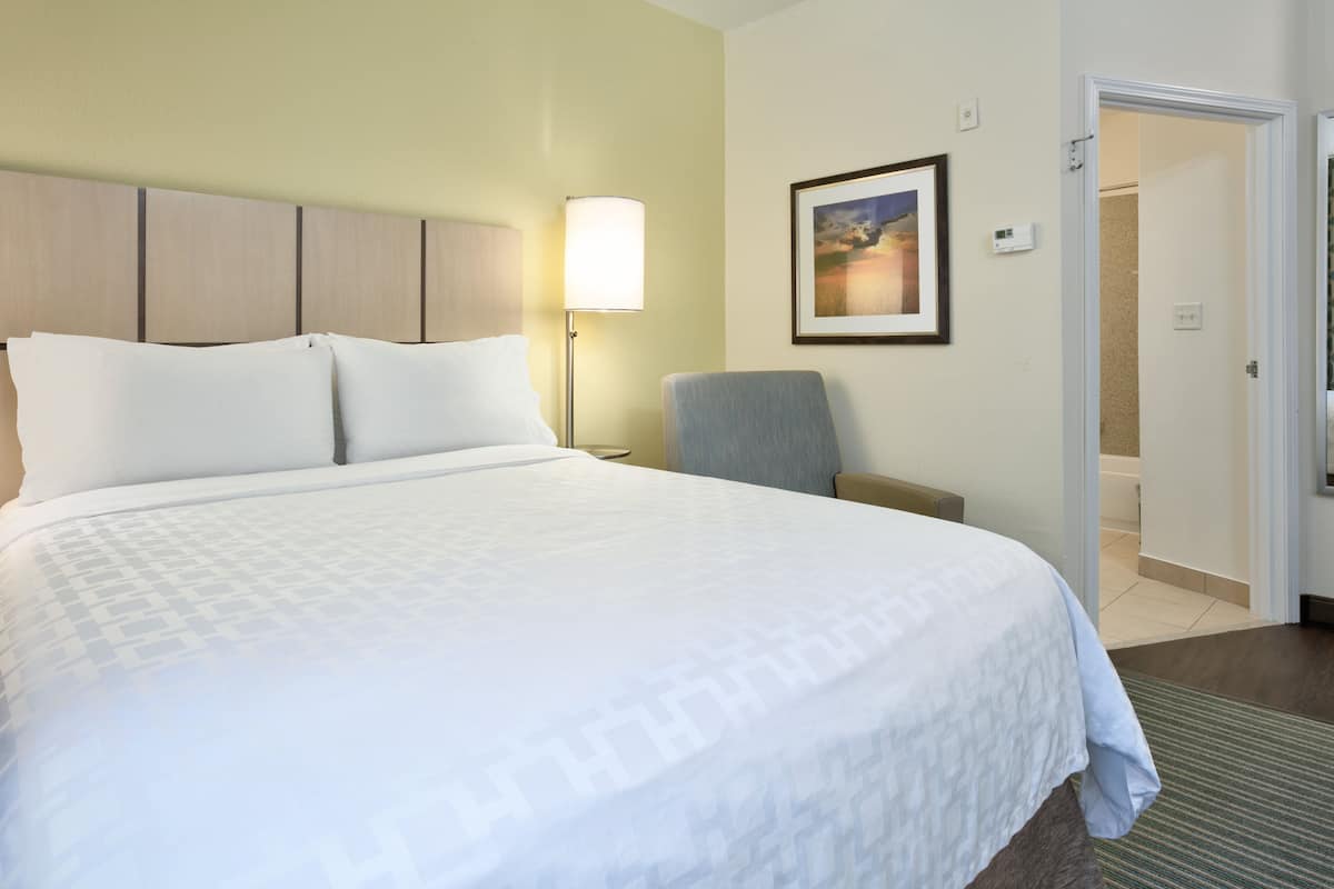 A hotel room with a large bed, a chair, a nightstand, a lamp, and a framed picture on the wall.