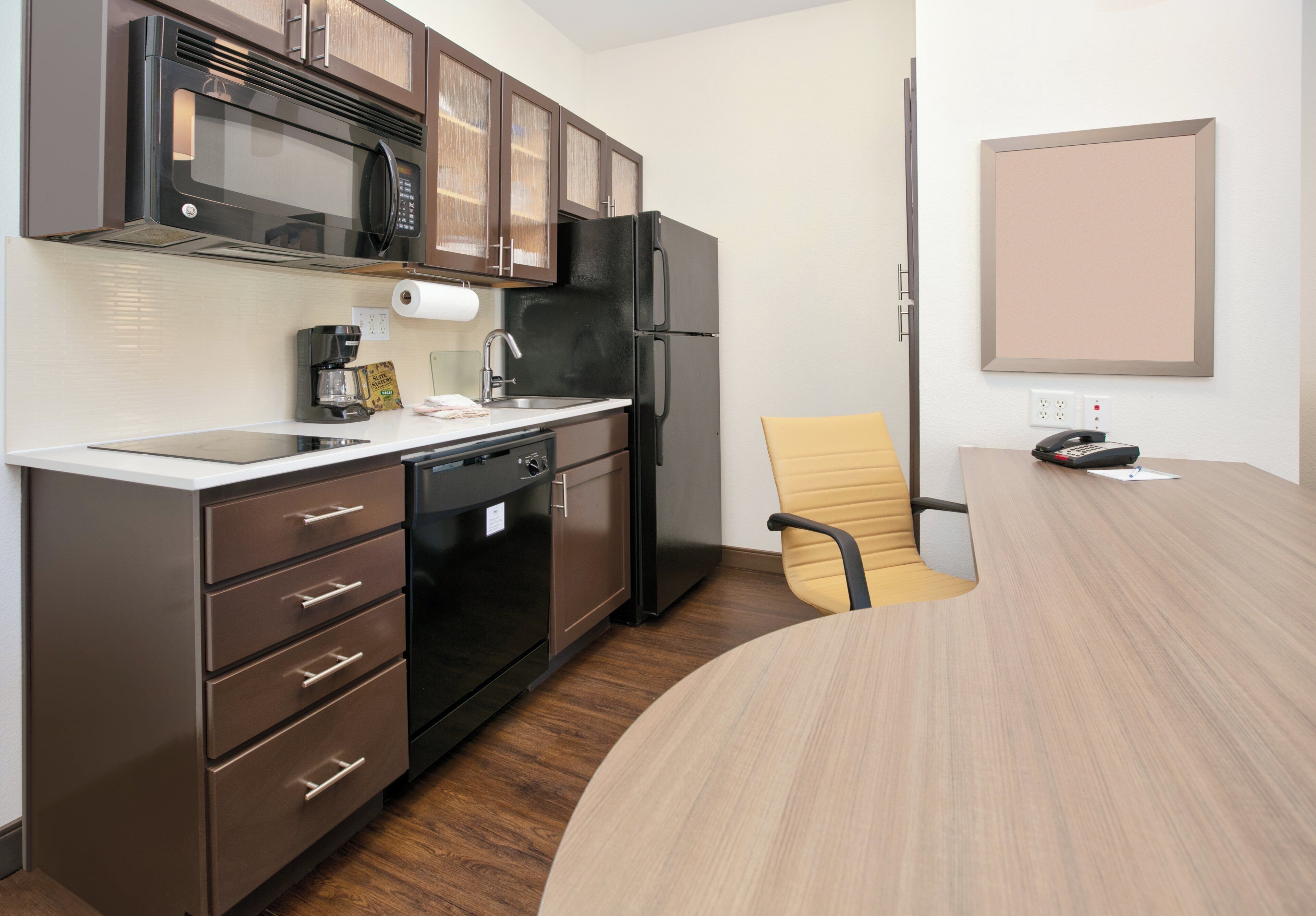 A compact kitchen with a microwave, coffee maker, and refrigerator. There's a wooden dining table with a chair.