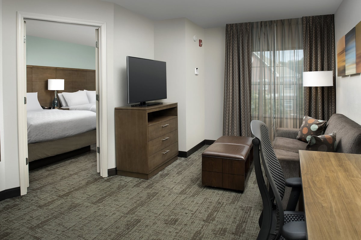 A hotel room with a bed, a TV on a wooden stand, a desk with a chair, and a window with curtains.