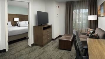 A hotel room with a bed, a TV on a wooden stand, a desk with a chair, and a window with curtains.