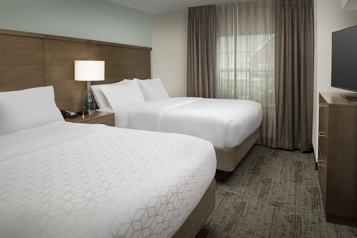 A hotel room with two beds, a nightstand with a lamp, a TV, and a large window with curtains.
