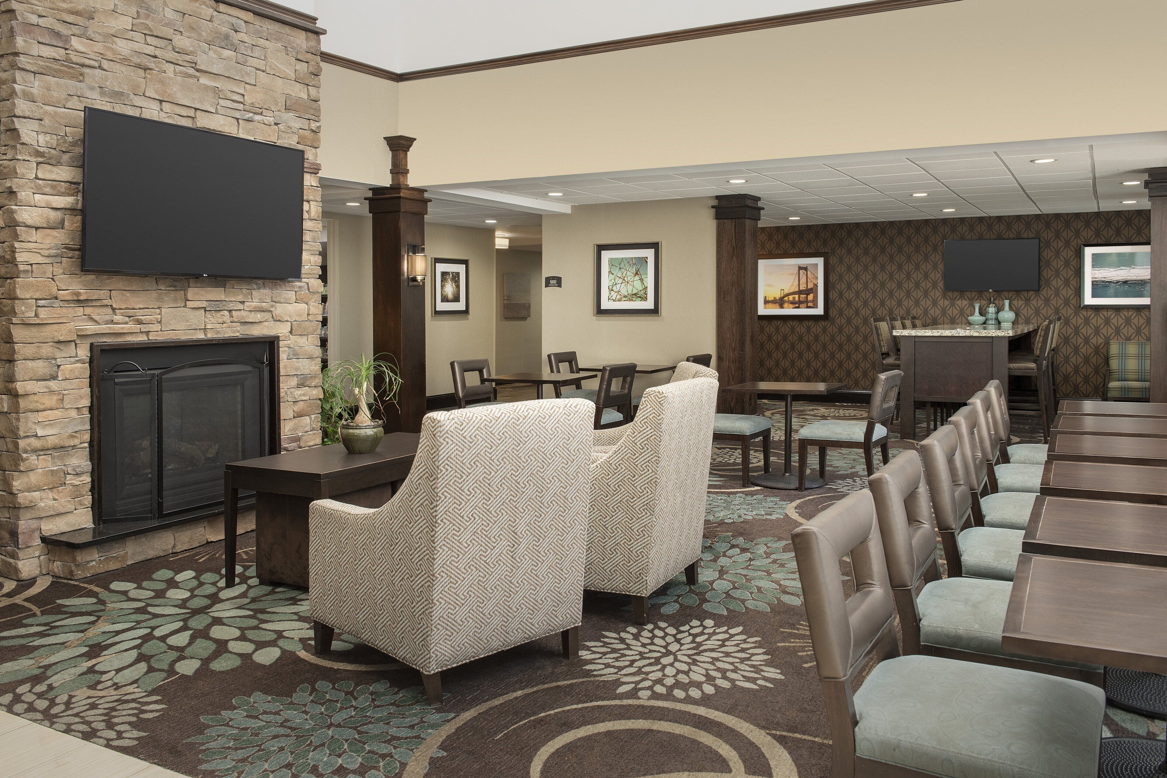 A hotel lobby with a stone fireplace, seating area with patterned chairs, a dining area with tables, and artwork on the walls.
