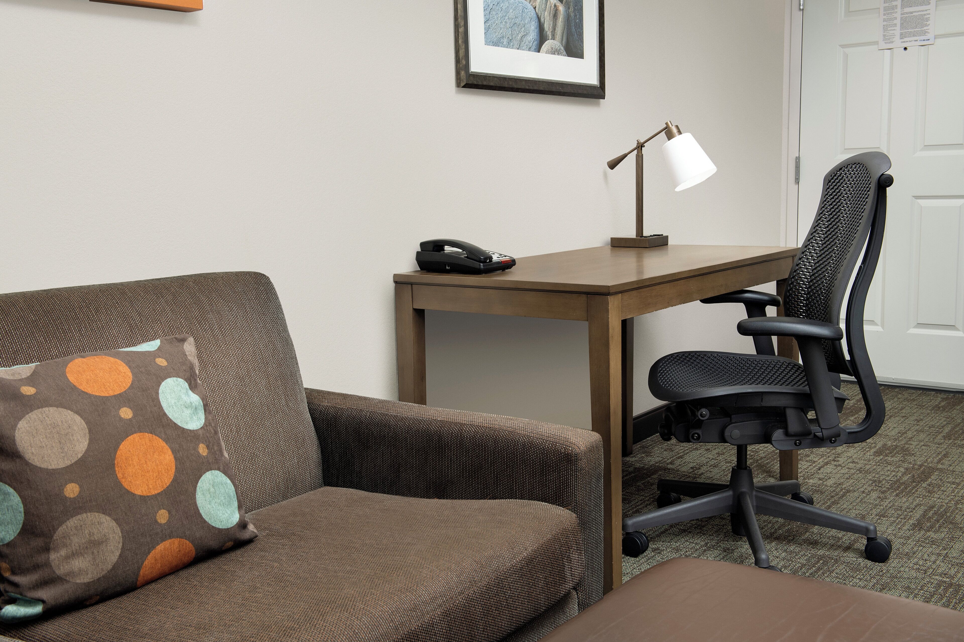 A hotel room with a brown sofa, a desk with a lamp, a black office chair, and a framed picture on the wall.