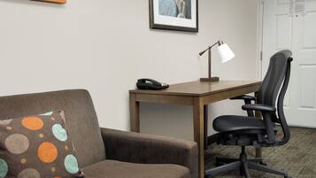 A hotel room with a brown sofa, a desk with a lamp, a black office chair, and a framed picture on the wall.