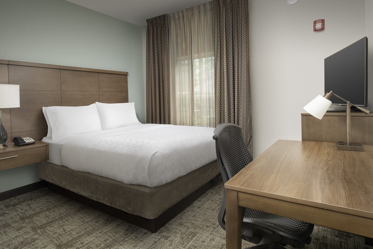 A hotel room with a bed, a desk, a chair, a lamp, a TV, and a telephone on the nightstand.