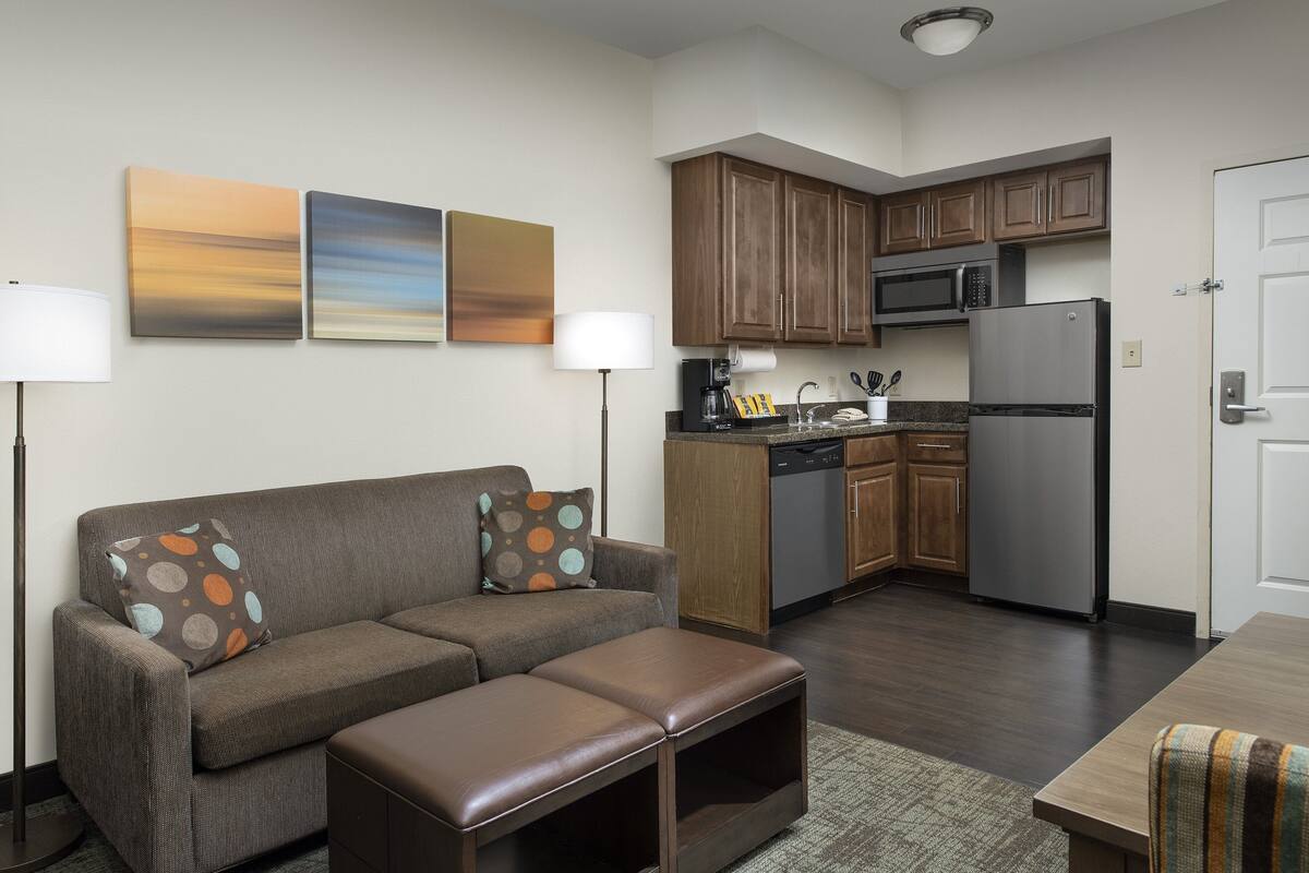 A modern living room with a sofa, ottoman, and a kitchen area with wooden cabinets and appliances.