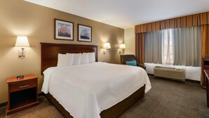 Hotelkamer met een groot bed, twee wandlampen, een nachtkastje met een klok, een stoel en een raam met gordijnen.