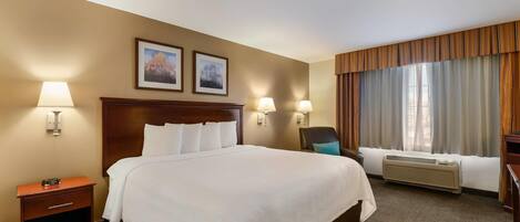A hotel room with a large bed, two wall-mounted lamps, a nightstand with a clock, a chair, and a window with curtains.