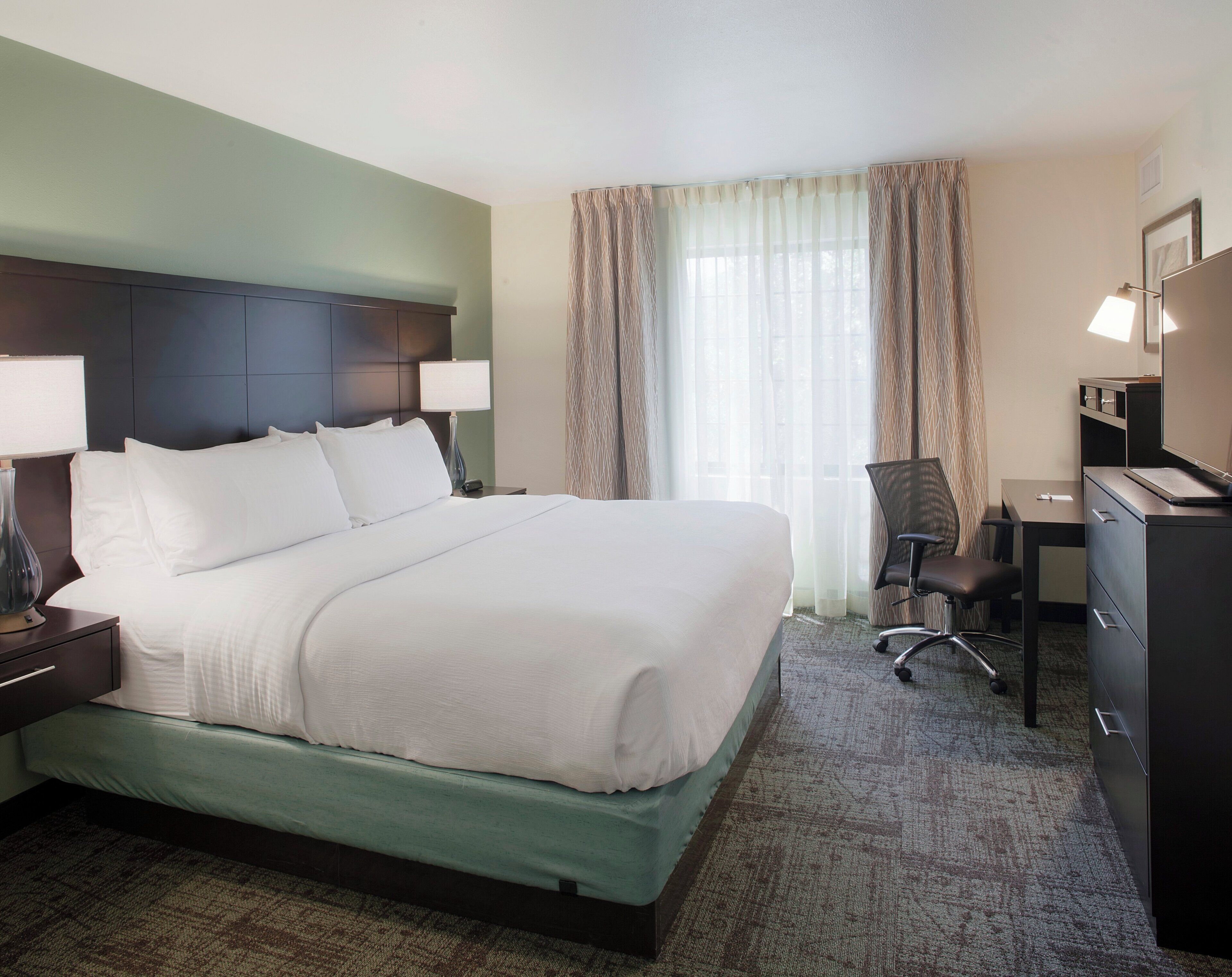 A hotel room with a large bed, a desk, a chair, and a dresser.