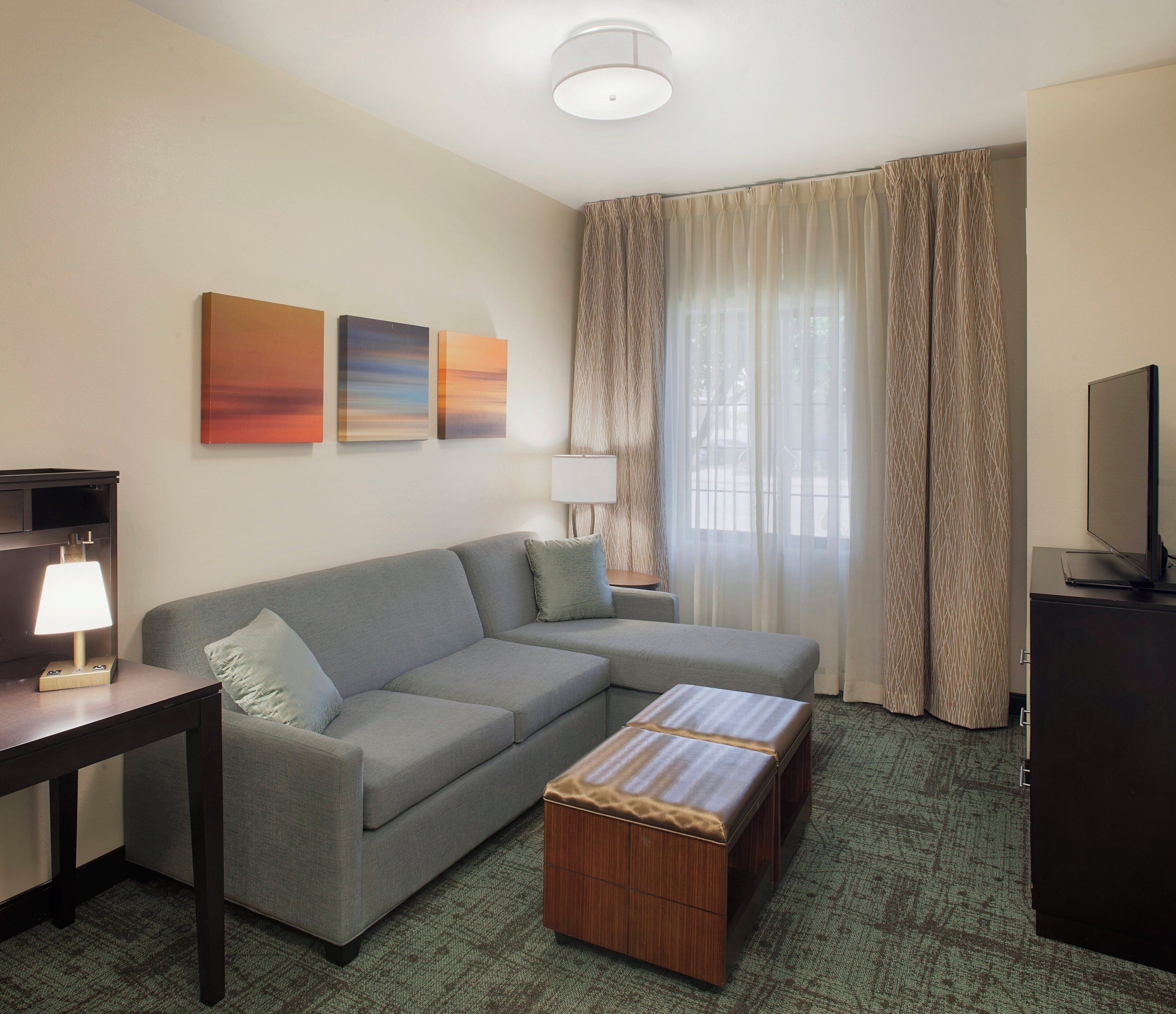 A modern hotel room with a grey sofa, a wooden ottoman, a flat-screen TV, and a wall-mounted lamp.