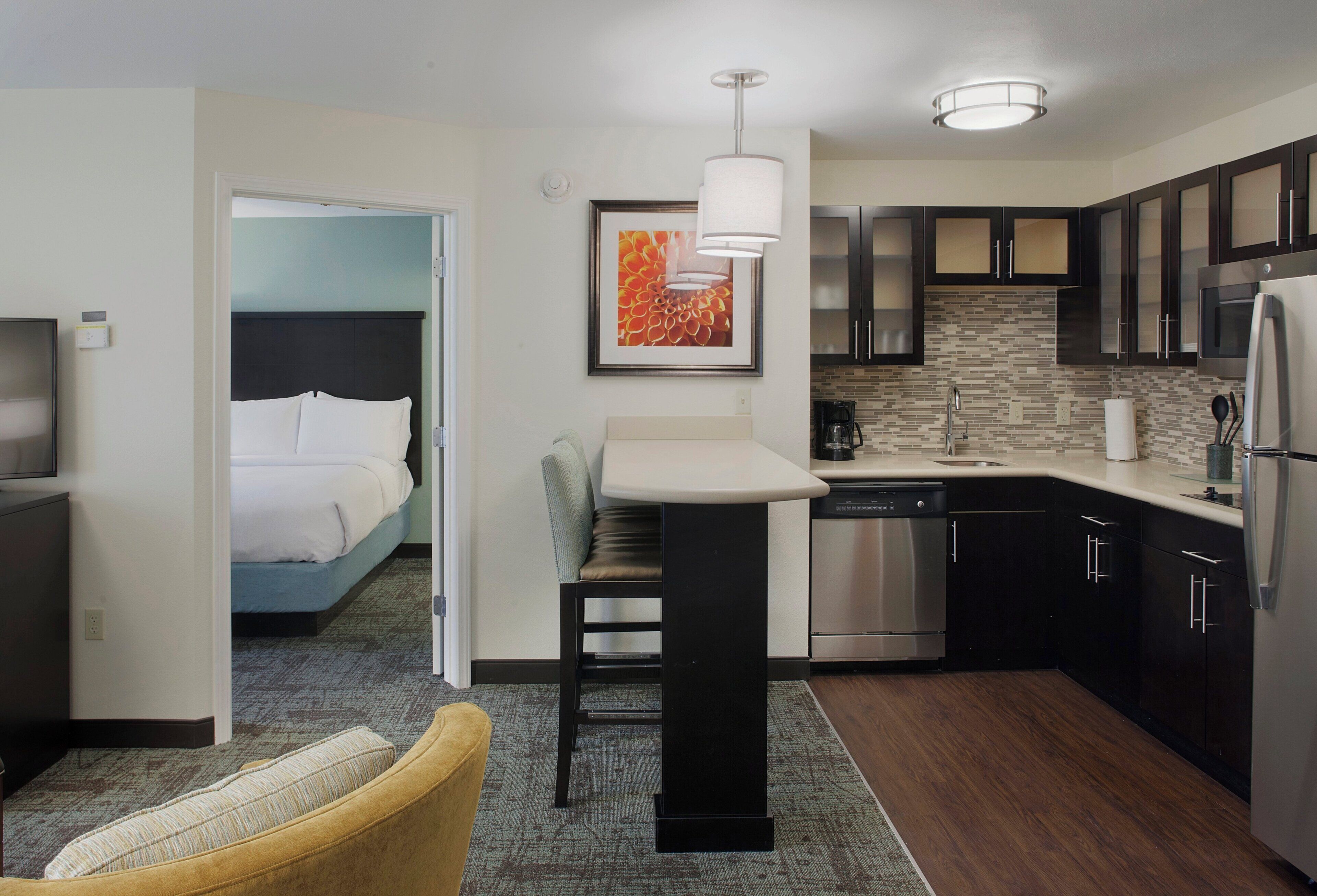 A compact hotel room with a bed, a small sitting area, a kitchenette, and a bathroom.