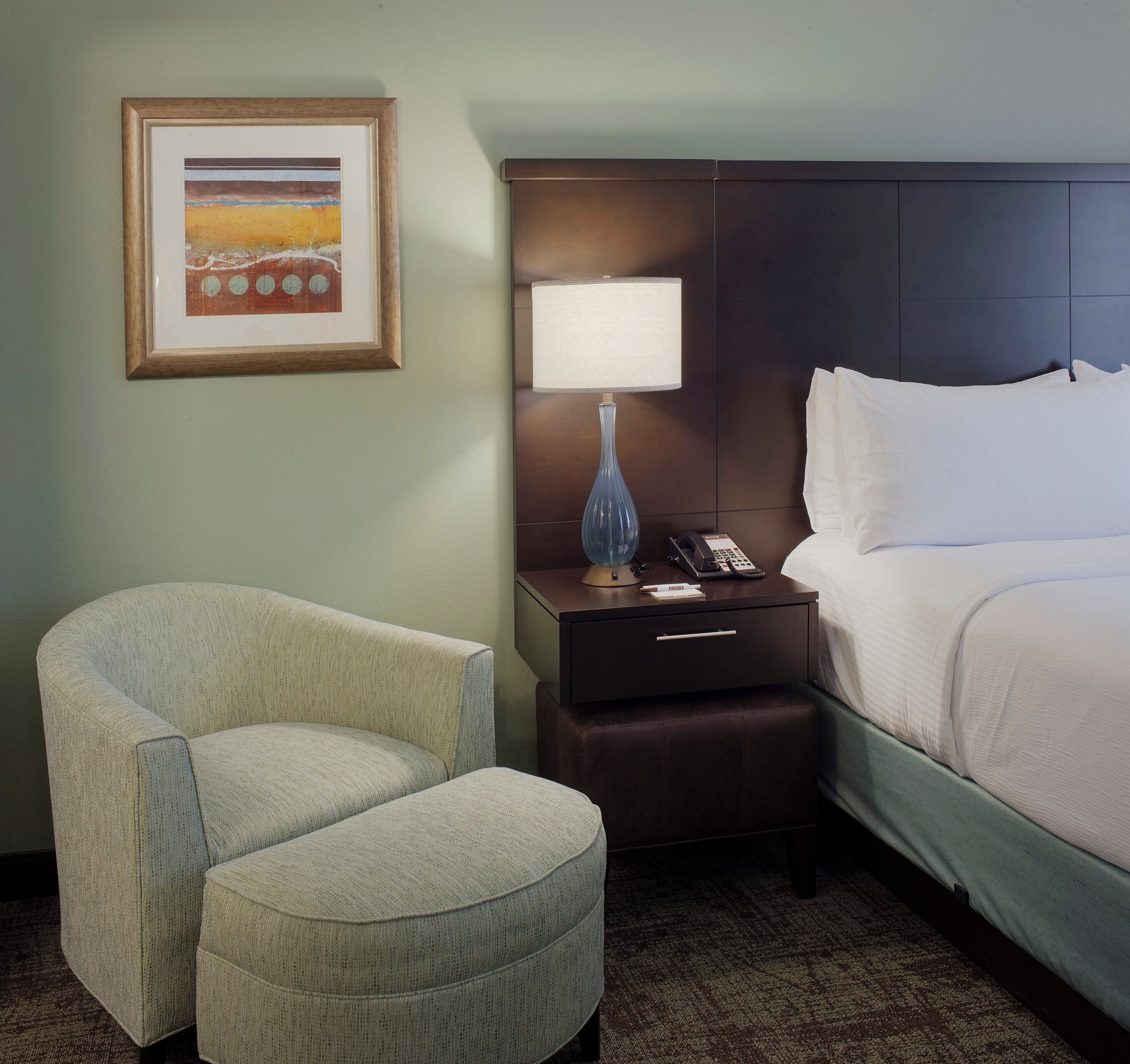 A hotel room with a bed, a chair, a nightstand with a lamp, a framed picture on the wall, and a carpeted floor.