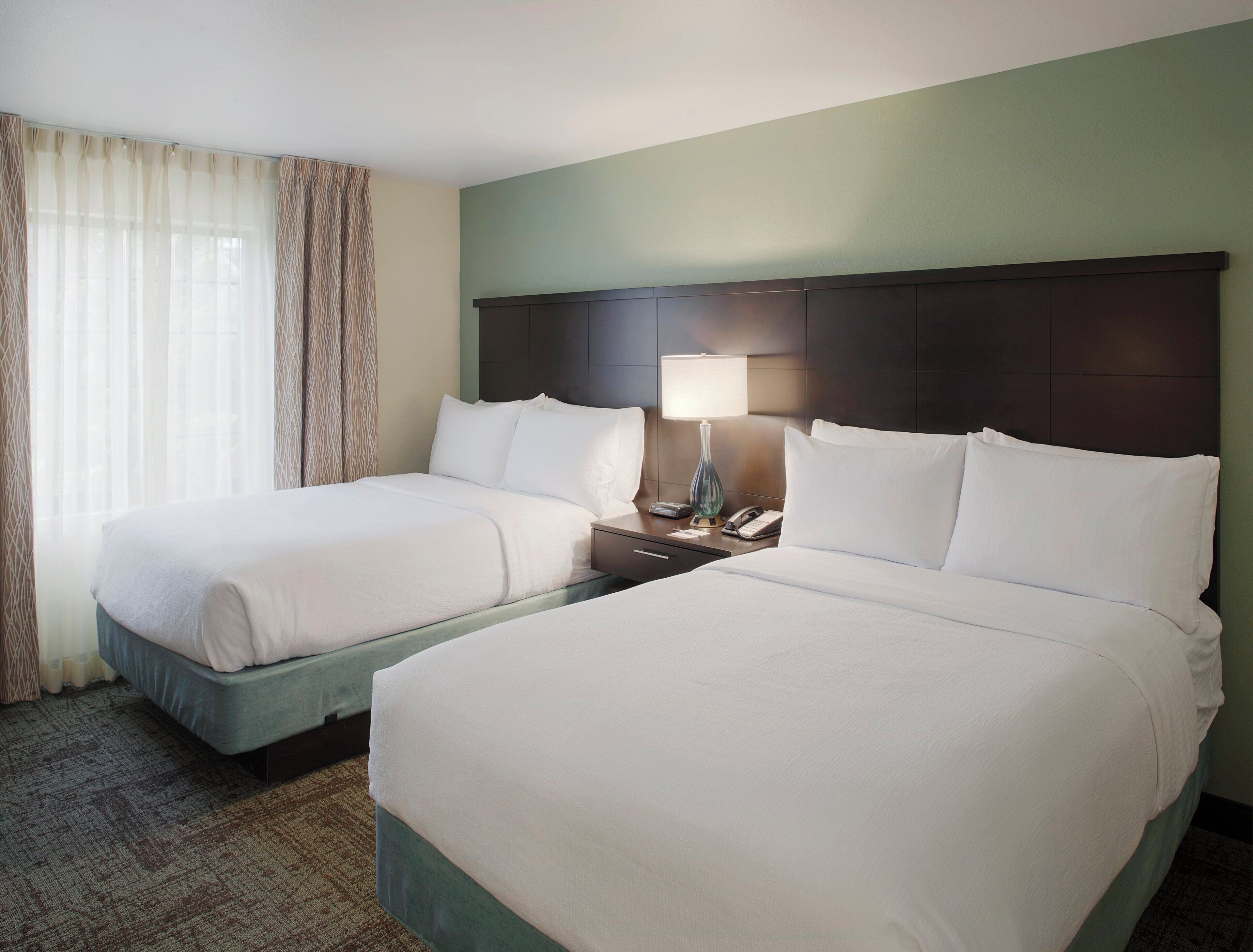 A hotel room with two beds, a nightstand with a lamp, and a window with curtains.