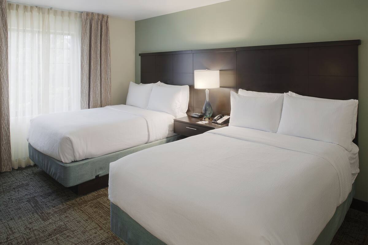 A hotel room with two beds, a nightstand with a lamp, and a window with sheer curtains.