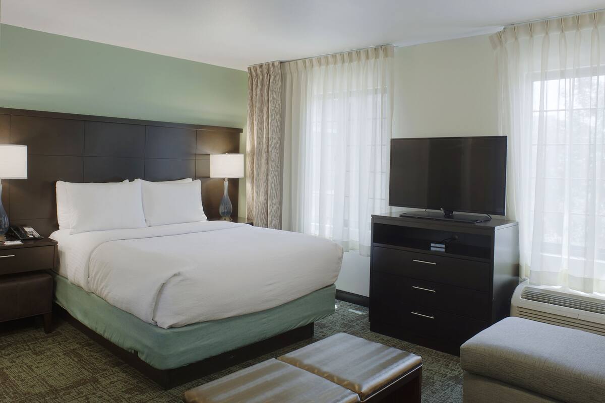 A hotel room with a large bed, a flat-screen TV, a desk with a lamp, and a window with sheer curtains.