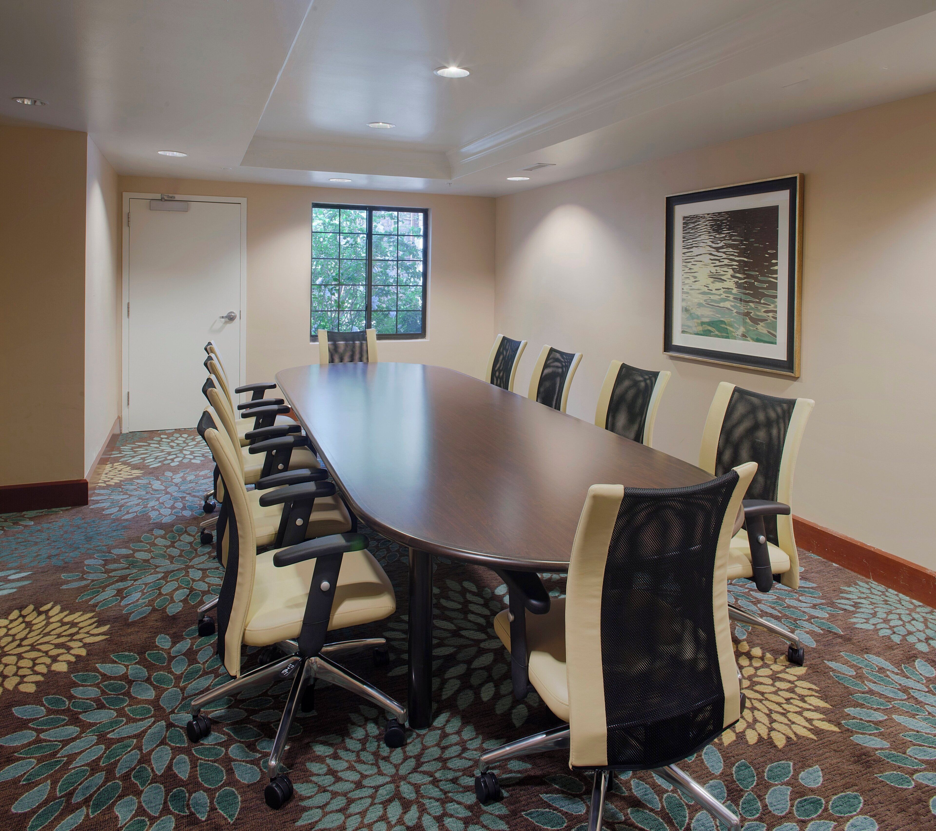 Un salón de conferencias con una gran mesa rodeada de sillas, una puerta, ventanas y un cuadro en la pared.
