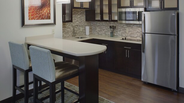 A modern kitchen with dark cabinets, a stainless steel refrigerator, a microwave, and a large island with bar stools. - Staybridge Suites Fayetteville/Univ Of Arkansas by IHG (Fayetteville)