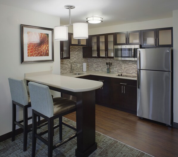 A modern kitchen with dark cabinets, a stainless steel refrigerator, a microwave, and a large island with bar stools. - Staybridge Suites Fayetteville/Univ Of Arkansas by IHG (Fayetteville)