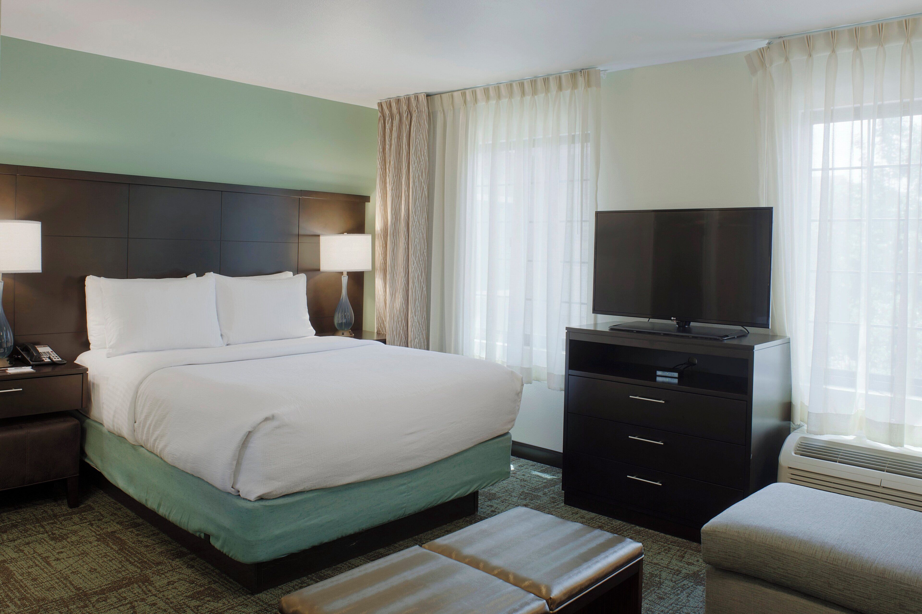 A hotel room with a bed, a desk with a lamp, a television on a dresser, and a window with curtains.